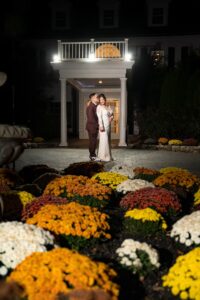 Mario and Idalia standing together in front of English Manor, illuminated by soft lighting, surrounded by vibrant fall flowers during their wedding celebration.
