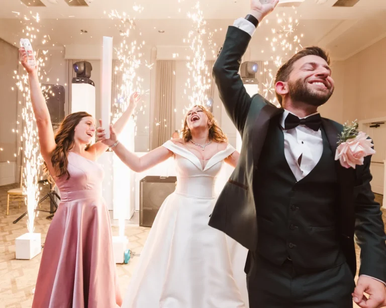 Groom in green tux raising hand in the foreground with a bride in a white wedding dress singing, looking up, and holding the outstretched arms of her bridesmaids dressed in pink dresses behind him, all in front of four floor sparks shooting off tall sparks with a white DJ setup  behind it.