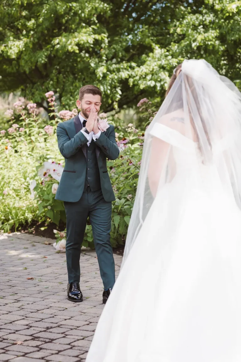 Happy groom with hands together up to mouth in the background about to open his eyes to see a bride in a white dress in the foreground