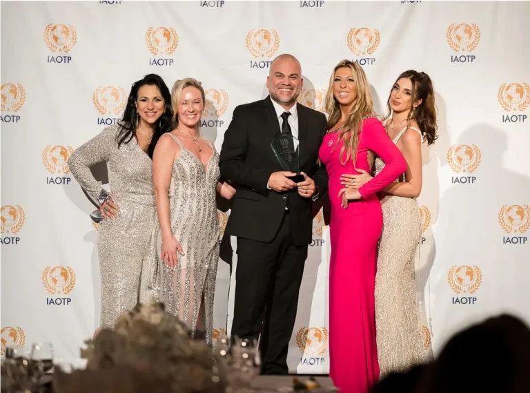 FNA Photographer Carlos Alvarado holding trophy by the IAOTP with 2 women on his left and 2 on his right all in front of the IAOTP step-and-repeat wall