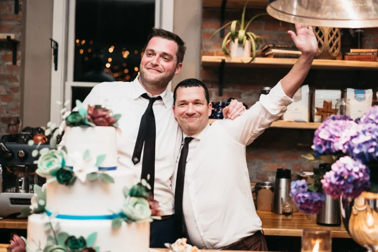 One groom with an arm around another groom whose arm is outstretched while behind a three-layer while cake with blue accents and flowers around, all in front of a window and bookshelf with pictures