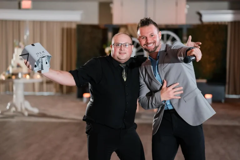An FNA DJ holding a white uplight with an arm around DJ Chris Hype who is pointing at the camera in a grey suit jacket