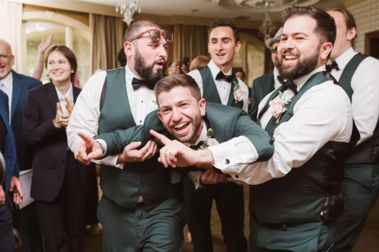 Smiling groom in a tux with with outstretched arms and pointed fingers being carried by four groomsmen in white button-downs, green vests and black bowties