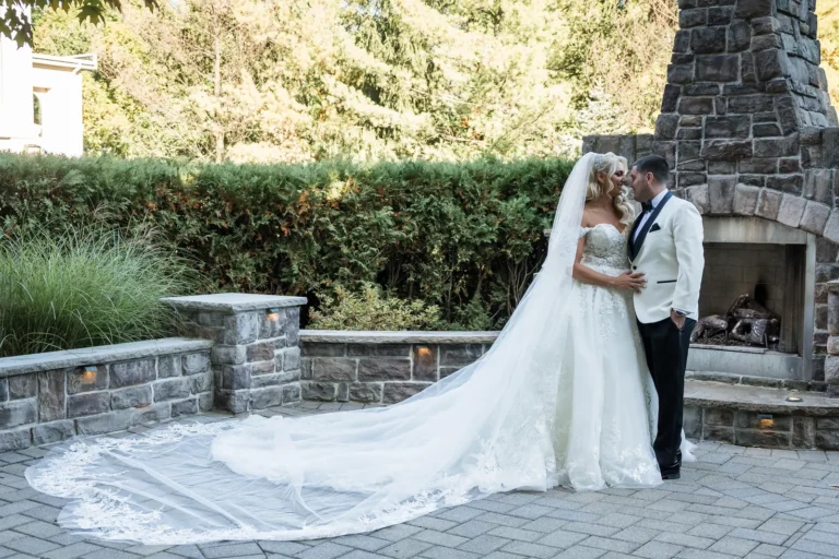 Bride in a white wedding dress with a long train next to her groom in a white tux with black accents, outside on a tiled patio with a fireplace, small wall, bushes behind it, and trees behind them.