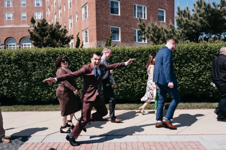 Groomsmen in a burgundy suit facing the camera while jumping sideways with outstretched arms in front of 2 other groomsmen in burgundy suits, behind a father in a blue suit next to his daughter in a flowery dress.