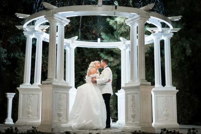 A bride in a white dress kissing her groom in a white tux while holding each other in the center of a circular white and stone gazebo all lit up from the inside.