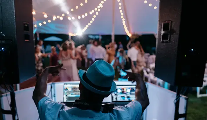 FNA DJ in a teal hat performing for a packed dance floor under a string-lit wedding tent